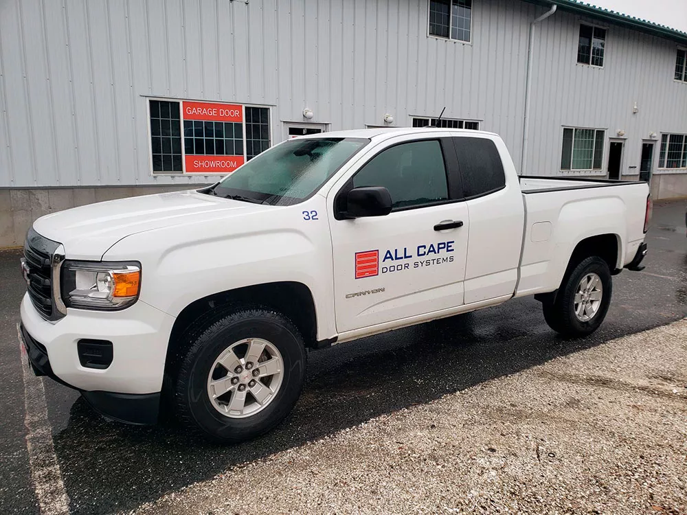 White pickup truck with All Cape Door Systems logo parked outside a building with Garage Door Showroom sign.